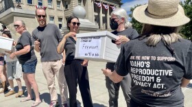 People lined up on the steps of the Idaho Capitol on a sunny day, handing each other white cardboard boxes. One woman with her back to the camera to the forefront right wears a t-shirt with the words "Ask me how to support Idaho's Reproductive Freedom and Privacy Act." She's just handed a box with the word "Fremont" sharpied on it to 