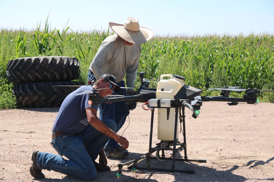 An older man in a wide brimmed, wicker sunhat and a grey long-sleeved shirt leans over a drone. It has black propellers that stick out and a cream colored tank in the middle. A younger man kneels next to it in a ballcap, sunglasses, a navy tee shirt and jeans. He is inspecting it. Behind them are two large tires stacked on top of each other and corn fields.