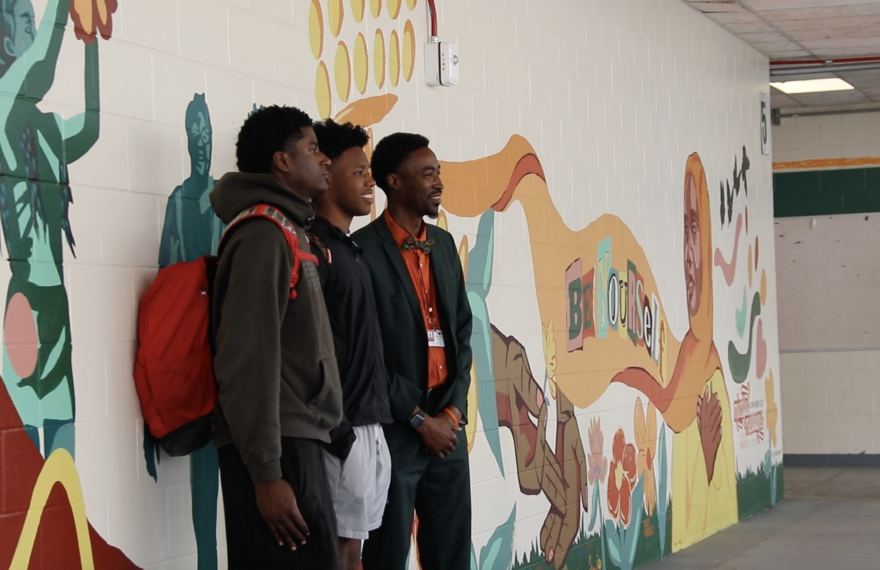 Two student artists pose on Tuesday, March 31, 2026, with Eastside Principal Leroy Williams in front of the section of the mural they completed. They painted themselves playing football. (Zoe Kahn/WUFT News) 