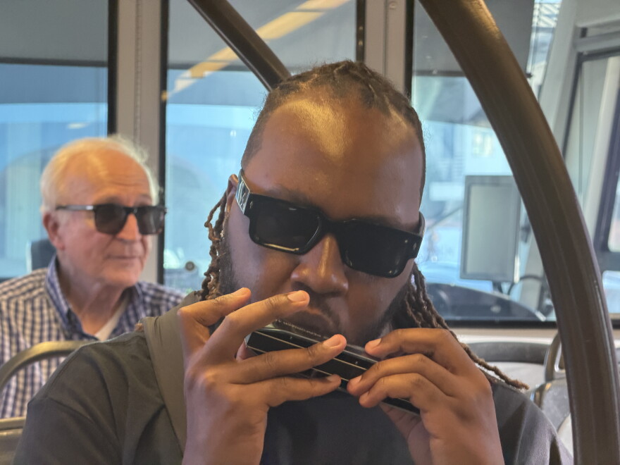 Man wearing sunglasses with long dreads plays a harmonica