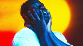 A Black man wearing a white t-shirt holds his face while standing in front of a colorful background