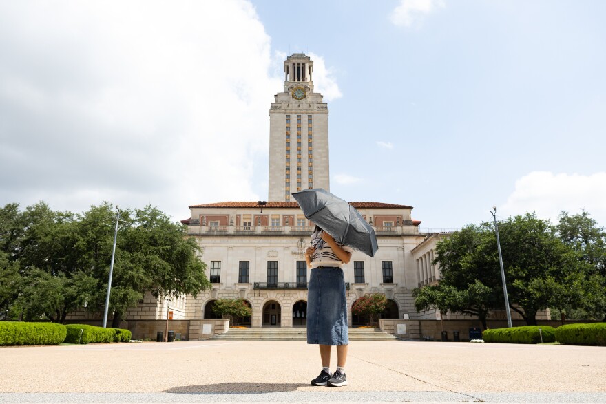A person holding an umbrella to cover her face stands in front of the UT Austin tower. 