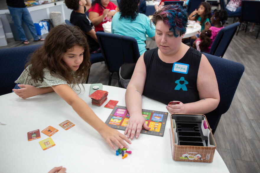 A young woman with short teal and magenta hair organizes cards for "Sushi Go" into a pile. A young girl with long hair reaches across the table toward them.