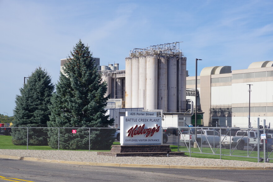 A white sign sits in front of a long metal fence topped with barbed wire. The sigh is for the Kellogg Battle Creek Plant, which can be partially seen in the behind the fence.