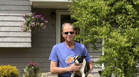 A man in a blue shirt and jeans stands in the grass, holding a small dog.