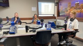 Three people sit at a a table in a radio talk studio: the man on the left has short grey hair and is wearing a blue short sleeved shirt; the woman in the center has short grey hair and is wearing a multi-colored shirt; the woman on the right has long red hair and is wearing a white t-shirt, jeans and sneakers.