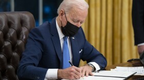 President Joe Biden sits at his desk and signs a document with a pen while wearing a black face mask.