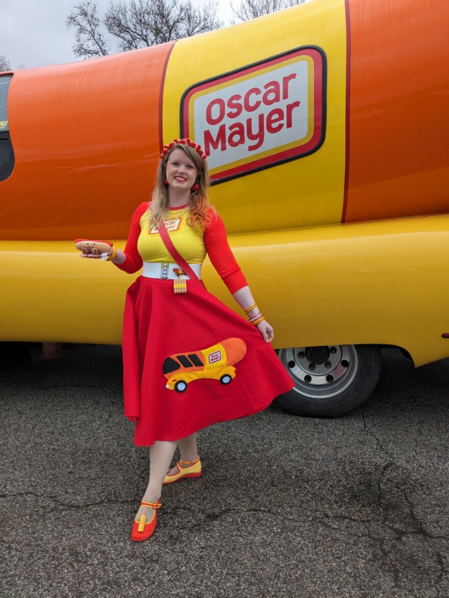 Amber Johnson stands in front of the Oscar Mayer Wienermobile in a coordinated outfit with a Wienermobile on her dress.
