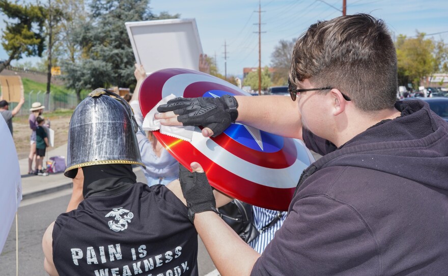 One protester wipes a red, white and blue Captain America–style shield held by another person wearing a helmet during a street protest, with other demonstrators visible in the background.