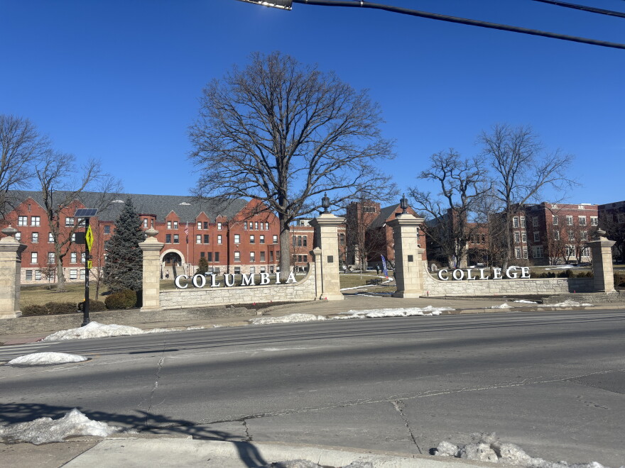 Patches of snow cover the ground in front of the gates of Columbia College on a sunny winter day. The word "Columbia" sits on the left gate and "College" on the right with a sidewalk in the middle. 