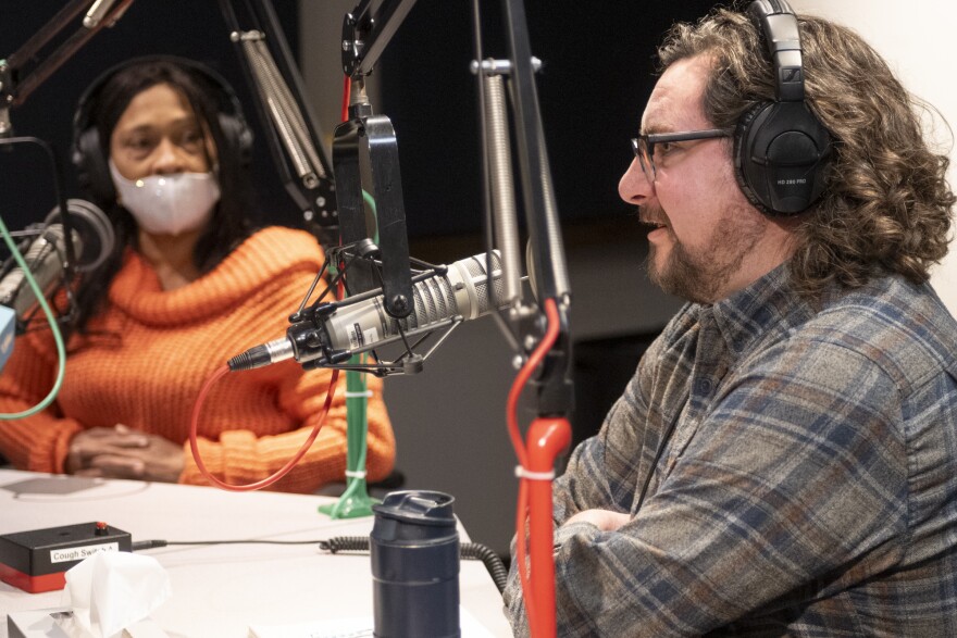 Two people sitting inside a radio studio are sitting in a radio studio. The man at right, Tom Meyer, is talking while Lisa Ray, left, is listening.