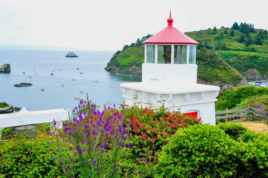 The Trinidad Memorial Lighthouse overlooks Trinidad Harbor on California’s North Coast, surrounded by green hills, colorful flowers and anchored boats in the Pacific Ocean.