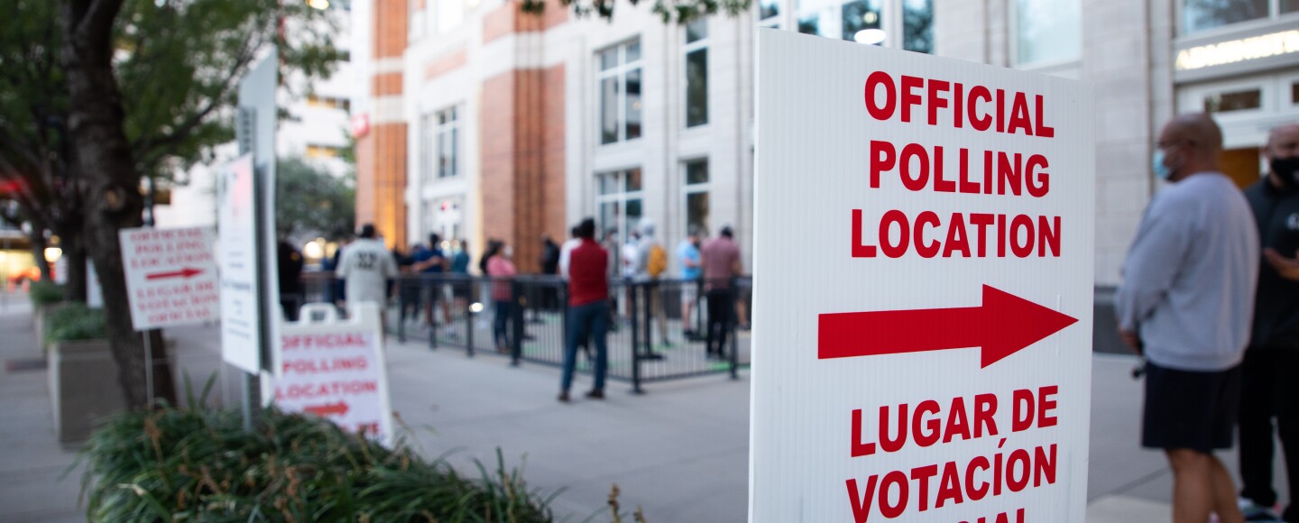 A polling location sign in English and Spanish sits in the forefront as voters wait in line.