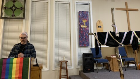 A person in a flannel and trans-themed head wrap stands at a church pulpit decorated with the gay flag. A nearby wooden cross is draped with a black scarf and the trans and nonbinary flags.