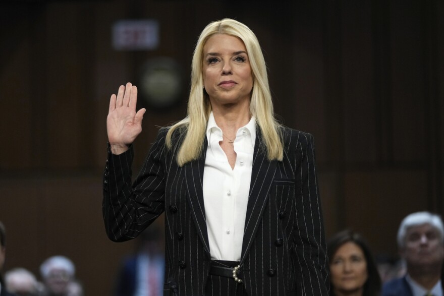 A woman in a dark jacket and white blouse and blond hair holds up her right hand as if she is being sworn in