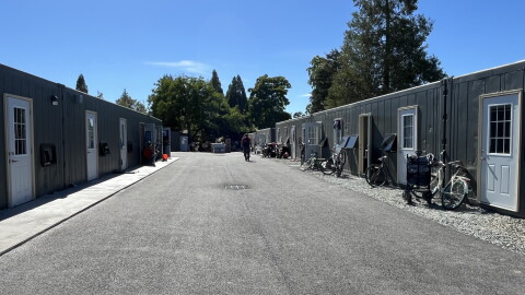 A wide driveway is lined with single-story shipping container homes. They are gray and have white doors and windows. 