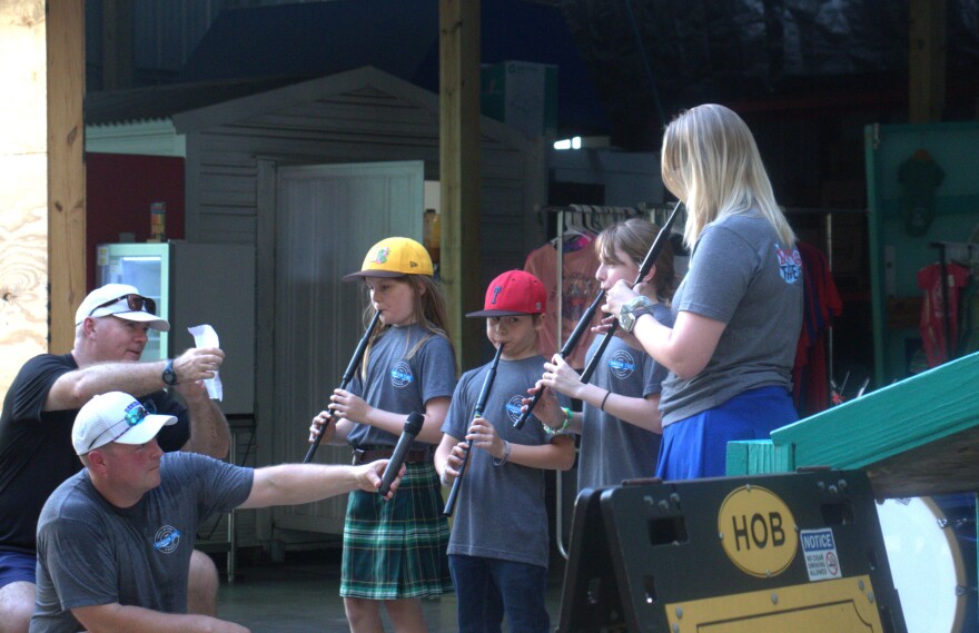 Three young kids, two of whom wear baseball caps, play long, reed-like instruments on a patio. A tall woman accompanies them, and two older men hold microphones up to the instruments. The boy on the left wears a green plaid skirt. A rack of T-shirts and a public restroom are visible behind them.