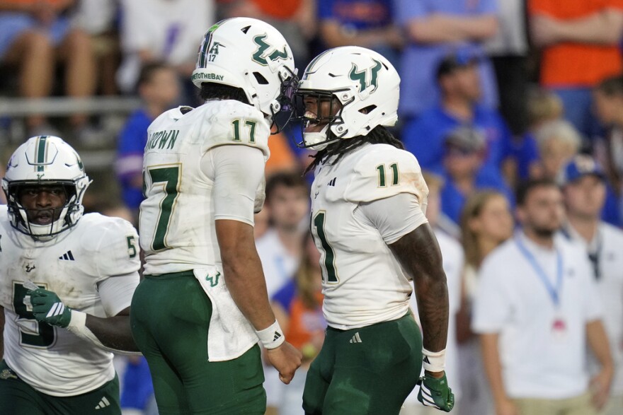 Two USF football players in white helmets and jerseys and green pants looking at each other celebrating