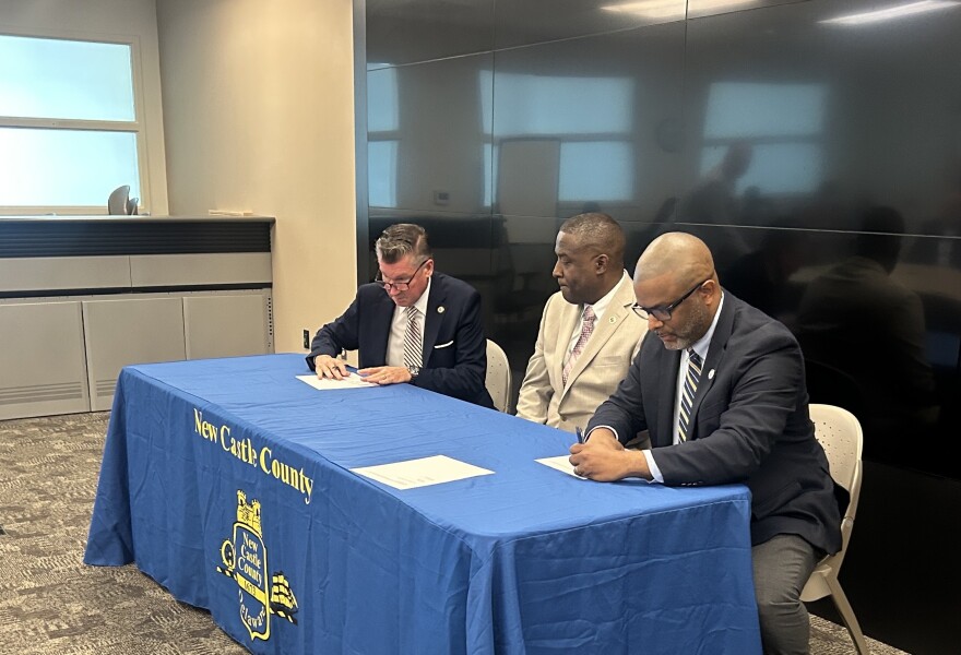 Three men sit at a table and sign papers. The table cloth has the New Castle County emblem. 