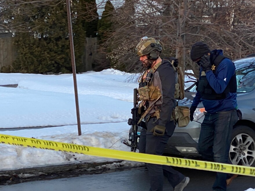 An armed FBI employee walks behind police tape, alongside a person on their phone, who appears to be wearing a police vest. There is snow and a car in the background. 