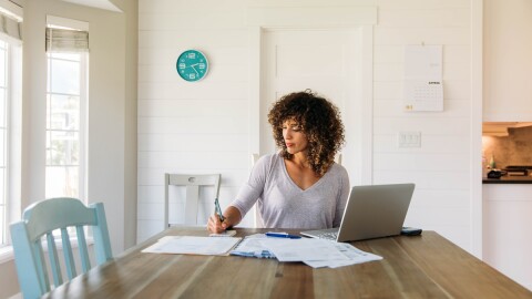 A woman sits at her dining room table with laptop and financial reports doing her monthly budget. She is writing down budgets as she works on her computer to do monthly finances, pay taxes and save money for the future.