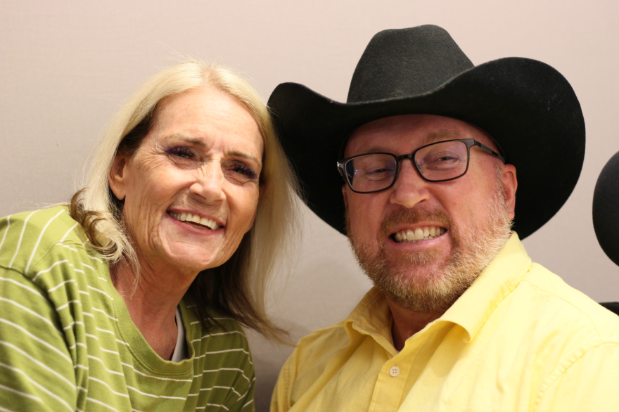 Kim Lake and her son Justin Tuft at their StoryCorps appointment in May 2025.  Kim has flipped, blonde, shoulder length hair.  She is 3/4 turned toward her son Justin.  She wears a green and white striped sweater.  Justin smiles, turned 3/4 toward his mother.  He has on a large black cowboy hat, black rimmed glasses, and a yellow button-up shirt.  The back and headrest of his wheelchair is partially visible behind him.