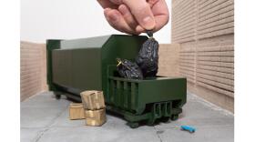 A hand dropping a miniature garbage bag into a miniature green dumpster