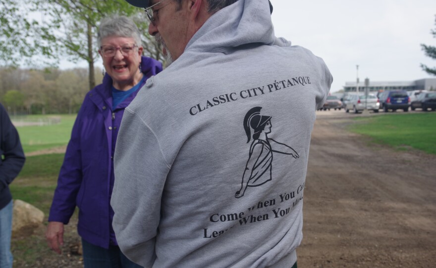 Man in grey sweatshirt faces away from camera. The back of the sweatshirt says "Classic city pétanque - come when you can, leave when you must" with image of person in ancient garb and helmet