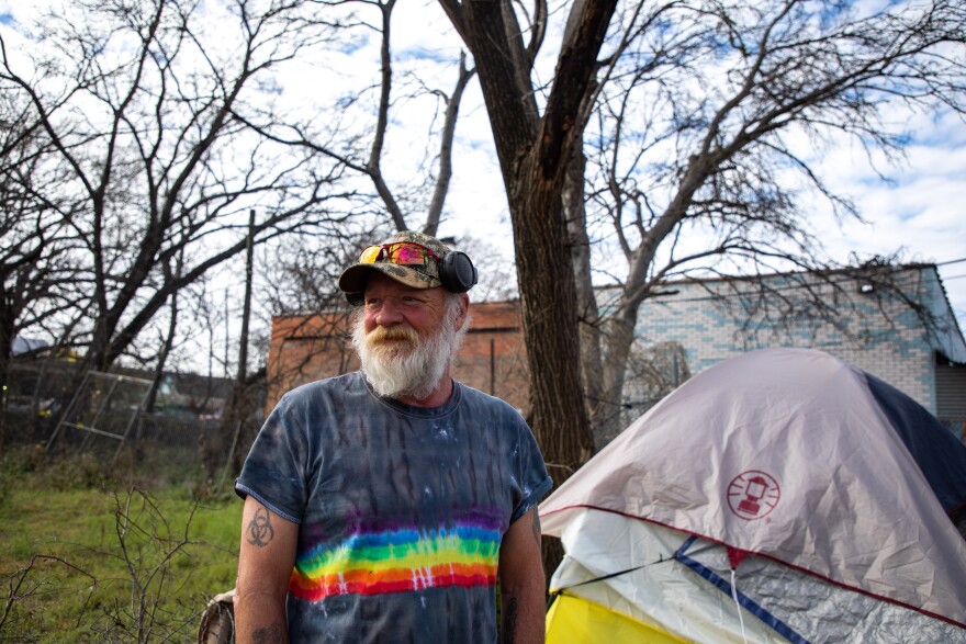 A man with a white beard stands in front of a tent and looks into the distance to the left. He's wearing a rainbow striped shirt, a green camouflaged hat and red tinted sunglasses on his hat.