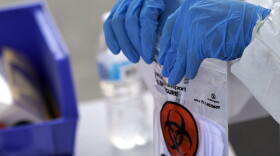 Blue-gloved hands hold a plastic bag with a red-and-black biohazard symbol on it. 