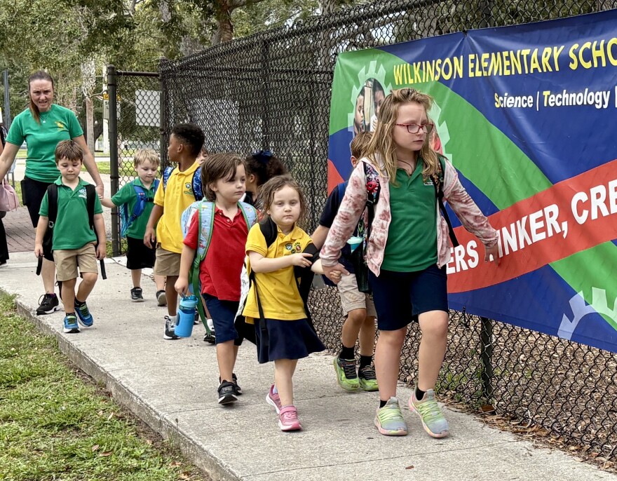 A group of young children walking next to a chain-link fence with an adult woman behind her