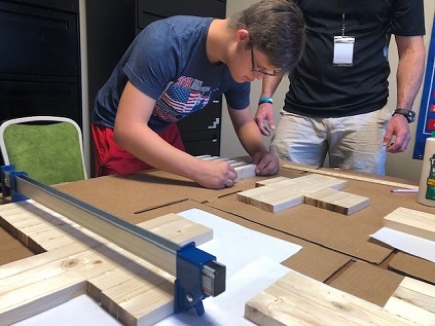 Kason Kolka is bent over a table next to David Kelly with various shapes of wood. He's helping create artwork for the VIP silent auction at The Producers concert later this month.