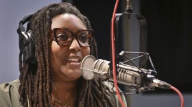 A person sits inside a radio studio talking at a microphone.