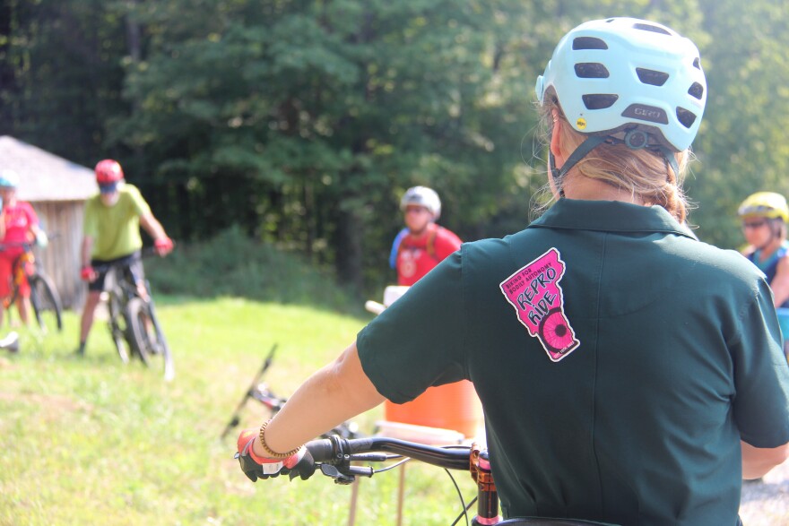 A woman wearing a black shirt and a blue helmet faces away from the camera holding the handle bars of a bike. The back of her shirt has a pink sticker in the shape of Vermont that says "Repro Ride"