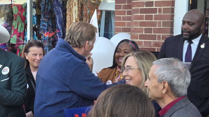 An outdoor campaign gathering where several people interact in front of a brick building. In the center, a man wearing a blue fleece jacket stands with his back partially to the camera, greeting and interacting with a smiling woman with blonde hair and glasses. Other smiling onlookers surround them. Some individuals in the crowd wear campaign buttons that read "Mike Rogers." In the background, a shop window displays colorful clothing, and white campaign balloons are partially visible.