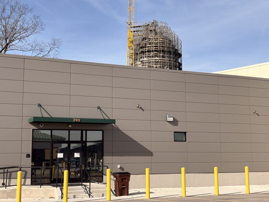 The temporary House and Senate chambers in Frankfort, Kentucky. The building's entrance as it faces the parking garage. The outside wall is made of flat, beige panels and has one window. There is a set of doors under a green awning, protected from car traffic by yellow bollards. The Capitol dome and a large crane is visible in the background, above the temporary building's roof.