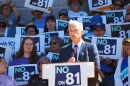 Denver District Attorney John Walsh speaks at a podium surrounded by people holding signs that say "No on 81"
