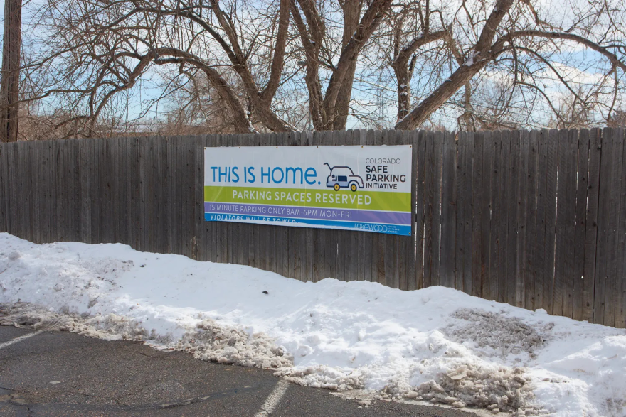 A parking lot with snow on the edges below a fence with a sign that reads "This is Home. Safe Parking Initiative" 