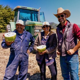 Three farmers stand in front of a tractor outside.