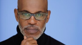 A Black man in a black turtleneck and glasses with a grey beard, looks toward the camera
