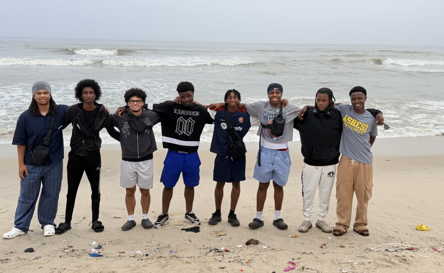 A group of young men with their arms around each other, standing on sand in front of a body of water