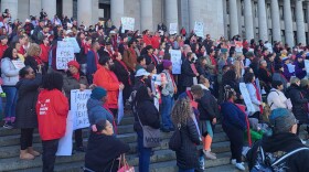 A crowd of people, some wearing red and holding signs in support of rent caps, are seen on the capitol steps in Olympia 