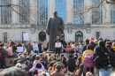 Hundreds of students gather by a large statue and an academic building. They hold signs, including one that reads "D-E-I or DIE" 