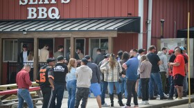A long ling of people stand outside a red, metal building. There are picnic tables outside and the sign on the building reads "Slap's BBQ."