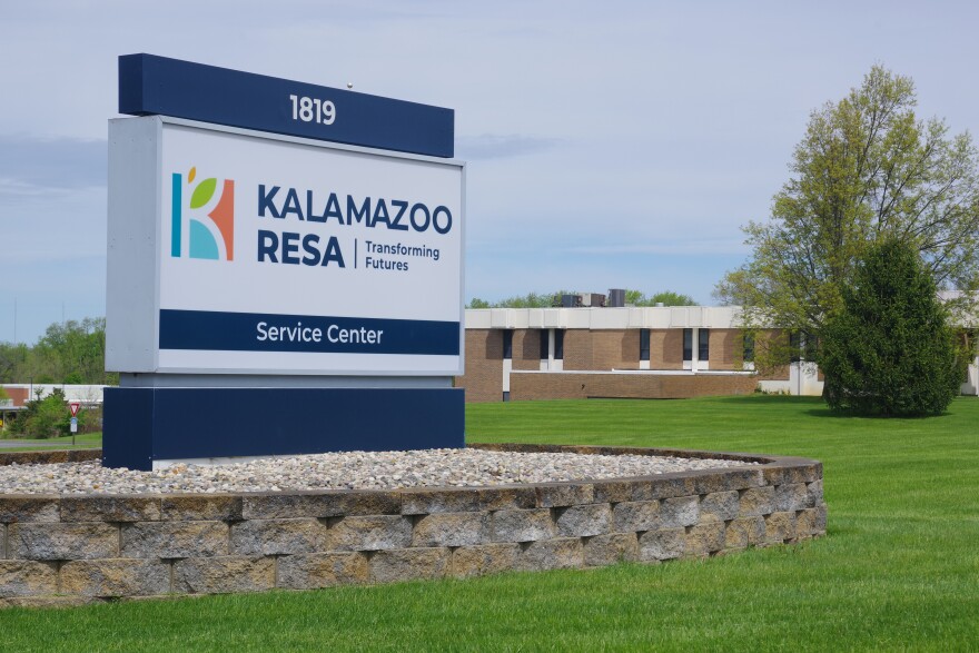 A white and blue sign sits at the left of the image, in the forefront of the picture. The sign reads "Kalamazoo RESA" with smaller text next to that reading "Transforming Futures." The sign sits in a platform of rocks, and encircled by a stone oval. Far back, beyond the green grass lawn sits a brown and white building.
