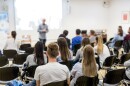 A speaker lectures in a university lecture hall as we see the backs of seated students listening