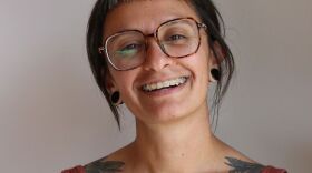 Dima Khalifa smiles. She has floral ttatoos across her chest, grazing her collarbone, and shoulders and wears an orange ribbed v-neck tank top.  