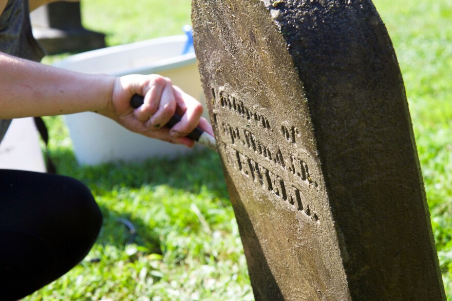 A photo of a gravestone and a hand holding a small brush on it, with grass in the background