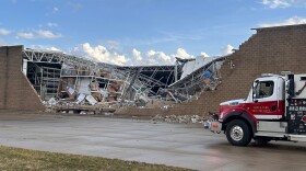 The side of a brown-red brick building is shown mostly collapsed, tall shelves on the inside and the roof have also caved in. The front of a firetruck can be seen on the pavement in front of the building. The vehicle sits at the middle right of the image