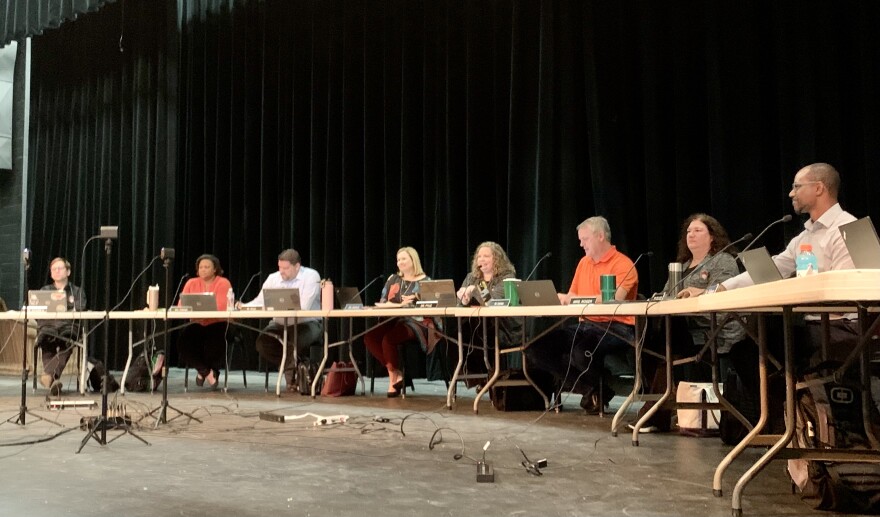 The Unit 5 school board sits at tables set on the stage of Normal Community West High School Auditorium. Each board member has a microphone, and a laptop in front of them. A black curtain hangs behind them onstage.